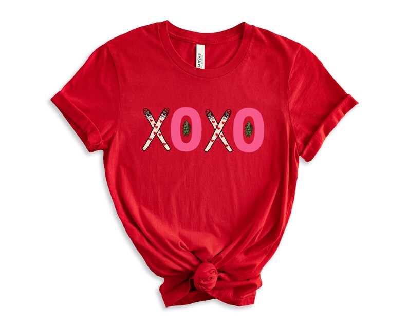May include: Red short-sleeved t-shirt with the word "XOXO" printed in pink letters. The letters are made up of crossed candy canes with red hearts and green Christmas trees.