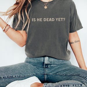 K&ouml;nnte beinhalten: Ein dunkelgraues T-Shirt mit dem Aufdruck "IS HE DEAD YET?" in hellbeige. Das Shirt wird von einer Person mit langen blonden Haaren und einer goldenen Halskette getragen.