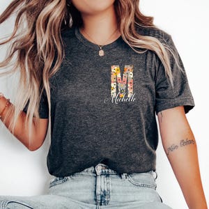 May include: A dark grey t-shirt with a floral letter "M" and the name "Michelle" printed on the front. The shirt is being worn by a person with long blonde hair.