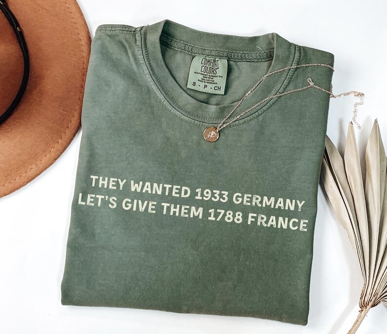May include: An olive green t-shirt with the text "THEY WANTED 1933 GERMANY LET'S GIVE THEM 1788 FRANCE" in white. A gold necklace with a pendant rests on the shirt. A brown hat and dried plant are in the background.