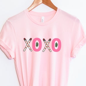 May include: Pink t-shirt with a graphic of the word "XOXO" made of crossed out cigarettes with hearts and a small pine tree.
