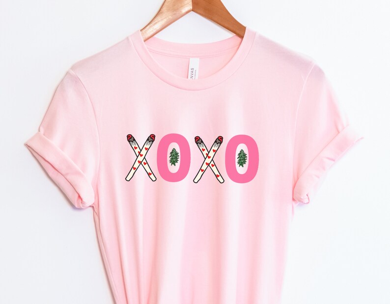 May include: Pink t-shirt with a graphic of the word "XOXO" made of crossed out cigarettes with hearts and a small pine tree.