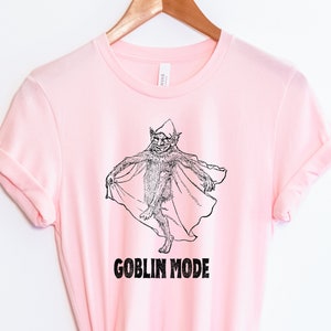 May include: Pink t-shirt featuring a black and white illustration of a goblin wearing a cape. The text "GOBLIN MODE" is printed below the illustration.