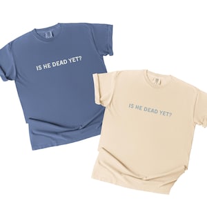 K&ouml;nnte beinhalten: Zwei T-Shirts mit dem Aufdruck "Is he dead yet?"  Ein T-Shirt ist hellblau und das andere beige.
