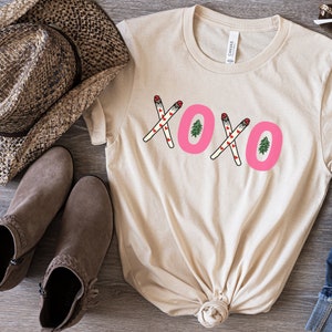 May include: A beige t-shirt with a pink "XOXO" graphic featuring small red hearts and green Christmas trees. The graphic is made up of two crossed sticks with red hearts and a green Christmas tree at the end of each stick.