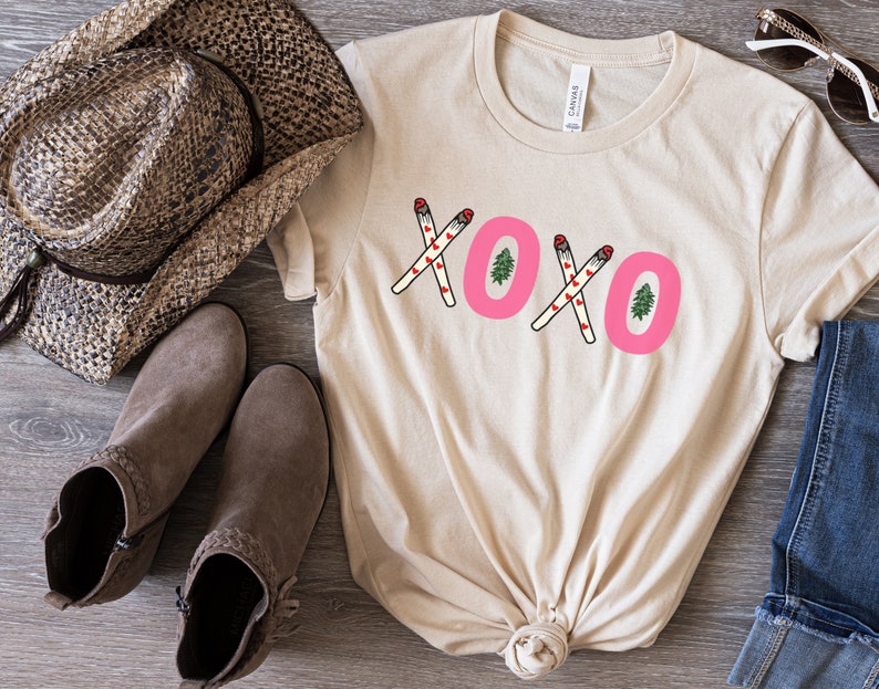 May include: A beige t-shirt with a pink "XOXO" graphic featuring small red hearts and green Christmas trees. The graphic is made up of two crossed sticks with red hearts and a green Christmas tree at the end of each stick.