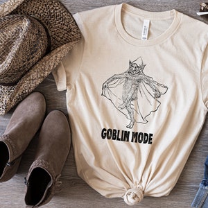 May include: A beige t-shirt featuring a black and white illustration of a goblin wearing a cape. The text "GOBLIN MODE" is printed beneath the illustration.