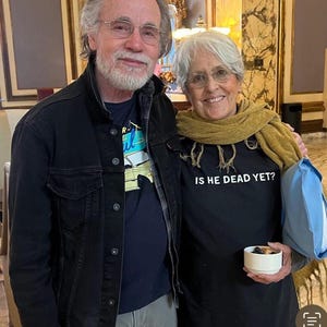 K&ouml;nnte beinhalten: Ein Mann und eine Frau stehen zusammen und l&auml;cheln. Der Mann tr&auml;gt eine schwarze Jacke &uuml;ber einem blauen Hemd mit einem Automotiv. Die Frau tr&auml;gt ein schwarzes T-Shirt mit dem Text "Is he dead yet?" und einen braunen Schal.