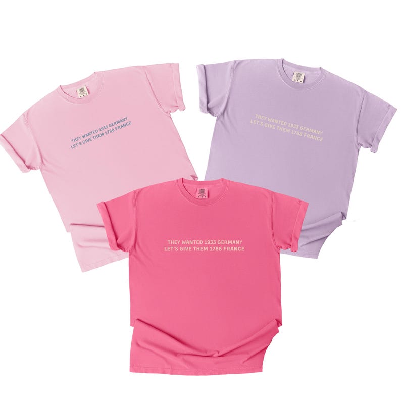 May include: Three pastel-colored t-shirts: light pink, lavender, and hot pink. Each shirt has the text "THEY WANTED 1933 GERMANY LET'S GIVE THEM 1788 FRANCE" printed on the front in a contrasting color. The shirts are made of soft material.