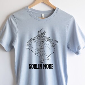 May include: Light blue t-shirt featuring a black and white illustration of a goblin wearing a pointed hat and a cape. The text "GOBLIN MODE" is printed below the illustration.
