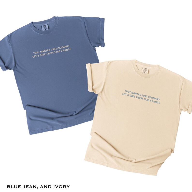 May include: Two short-sleeved t-shirts, one blue and one ivory, are displayed. Each shirt has the text "THEY WANTED 1933 GERMANY LET'S GIVE THEM 1788 FRANCE" printed in a light blue color. The shirts are made of a soft material.
