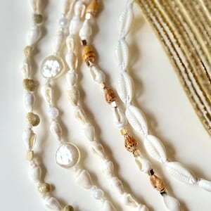 Peut inclure: Un collier multi-rangs composé de coquillages blancs, de perles en bois brun et de perles en métal doré. Le collier est superposé et a un style bohème.