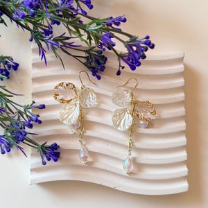 Peut inclure: Une paire de boucles d'oreilles pendantes dorées composées de breloques en forme de pétales translucides blancs et transparents, de petites perles sphériques blanches et de cristaux en forme de goutte irisés suspendus à des chaînes.