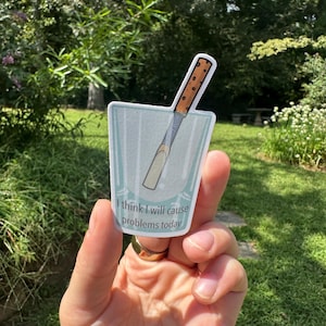 May include: A die-cut sticker featuring a light blue glass containing a brown and tan wooden reed, with the text "I think I will cause problems today" printed in black on the front of the glass, showcasing a humorous and illustrative design.