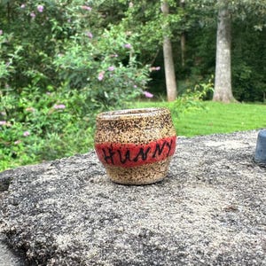May include: A small, round ceramic pot with a speckled tan and brown finish. A horizontal red band wraps around the center, featuring the word "HUNNY" written in black, hand-painted, cursive lettering. The pot has a wide opening and a tapered base.