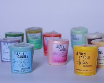 May include: A collection of scented votive candles in various colors, including blue, pink, yellow, and green. Each candle has a label with the brand name "Eleni's Candle" and a scent description. The candles are hand-poured and made with a soy blend.