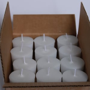 May include: A cardboard box filled with white unscented votive candles. The candles are arranged in rows and are ready to be used.