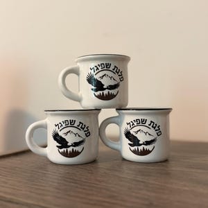 May include: Three white ceramic mugs with black rims, stacked in a pyramid formation. Each mug features a black graphic of a flying eagle over mountain peaks and forest silhouettes, with Hebrew text arched above the eagle design.