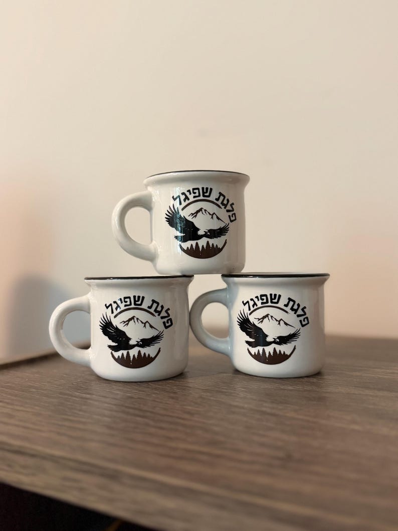 May include: Three white ceramic mugs with black rims, stacked in a pyramid formation. Each mug features a black graphic of a flying eagle over mountain peaks and forest silhouettes, with Hebrew text arched above the eagle design.