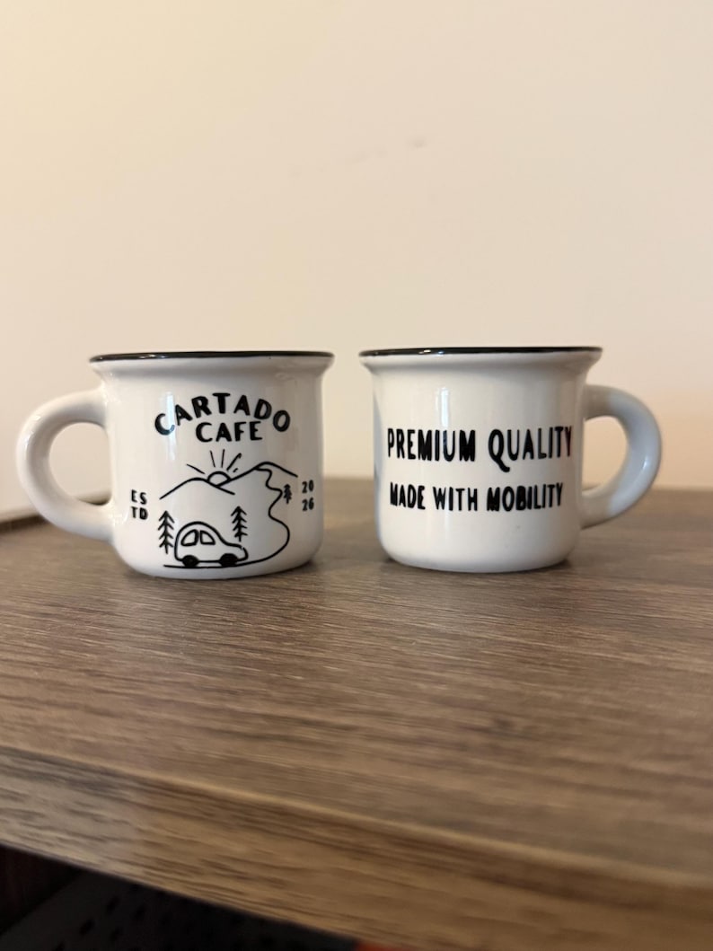 May include: Two white ceramic espresso mugs with black rims and handles. The left mug features a black graphic of a car, trees, and the text "CARTADO CAFE ESTD 2026". The right mug displays the black text "PREMIUM QUALITY MADE WITH MOBILITY".