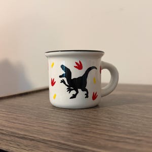 May include: A small white ceramic mug with a black rim and a handle. The exterior features a black silhouette of a dinosaur surrounded by small red dinosaur footprint icons and yellow dash marks scattered across the white surface of the mug.