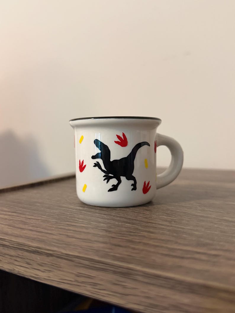 May include: A small white ceramic mug with a black rim and a handle. The exterior features a black silhouette of a dinosaur surrounded by small red dinosaur footprint icons and yellow dash marks scattered across the white surface of the mug.
