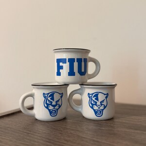 May include: Three white ceramic mugs with a black rim. The top mug features the blue text FIU. The two bottom mugs each display a blue graphic of a panther head. Each mug has a single handle on the side and a smooth, glossy finish throughout.