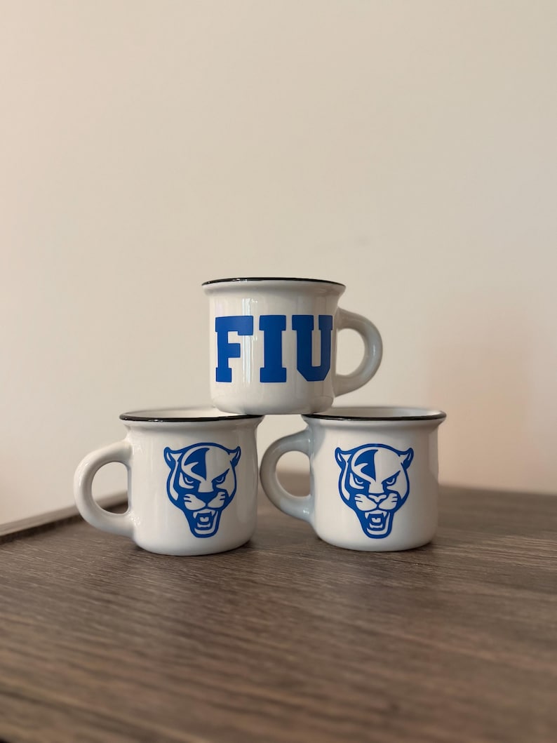 May include: Three white ceramic mugs with a black rim. The top mug features the blue text FIU. The two bottom mugs each display a blue graphic of a panther head. Each mug has a single handle on the side and a smooth, glossy finish throughout.