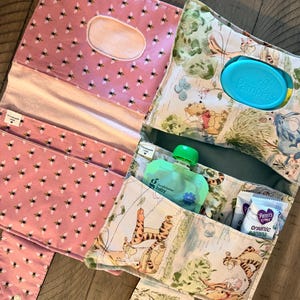 May include: A handmade baby changing station with a pink bee print and a Winnie the Pooh print. The station includes pockets for wipes, snacks, and baby food. A teal Pampers wipes container is visible.