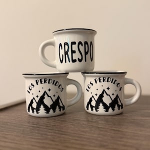 May include: Three white ceramic mugs with black rims and handles. The top mug features the text CRESPO in black lettering. The two bottom mugs feature a black graphic of mountains, pine trees, and stars with the text LOS PERDIDOS above them.
