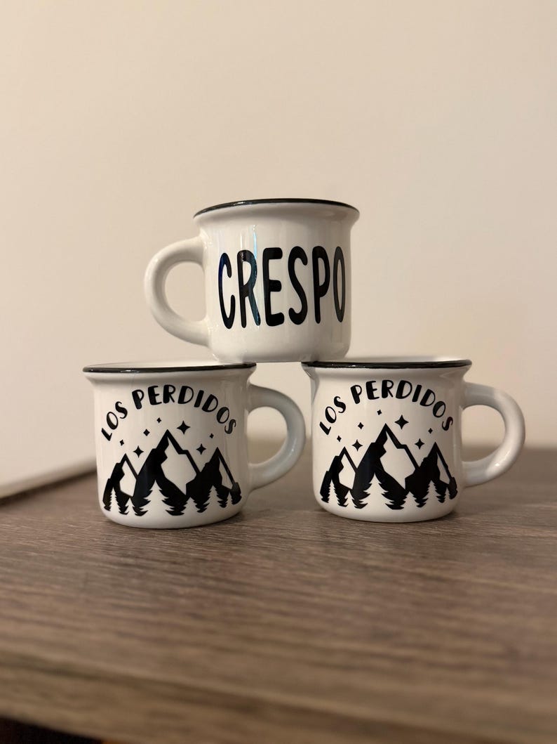 May include: Three white ceramic mugs with black rims and handles. The top mug features the text CRESPO in black lettering. The two bottom mugs feature a black graphic of mountains, pine trees, and stars with the text LOS PERDIDOS above them.