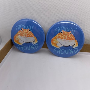 May include: Two blue and white round buttons with a cartoon orange frog on each. The frog is sitting on a blue background with the text "Froggin' Around" in white.