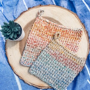 May include: Two crocheted pot holders in a rainbow colour pattern. The pot holders are on a wooden surface.