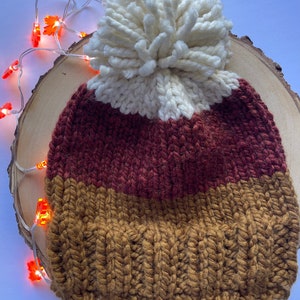 May include: A knitted beanie with a white pom pom. The beanie is striped with brown, rust, and white.