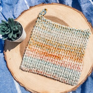 May include: A crocheted pot holder with a hanging loop. The pot holder is made with a multi-coloured yarn in shades of green, orange, and beige.