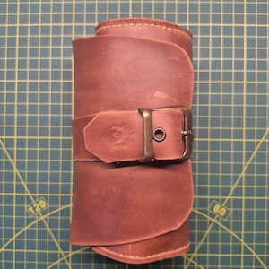 May include: Rolled brown leather item secured with a buckle. The leather appears distressed with visible stitching. The buckle is a dark metal color. The item is rolled up and secured with a strap and buckle.