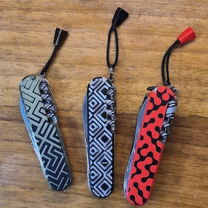 May include: Three multi-tool pocket knives with patterned designs. One has a black and gold geometric pattern, another has a black and white diamond pattern, and the third has a red and black abstract design. Each knife has a lanyard attached.