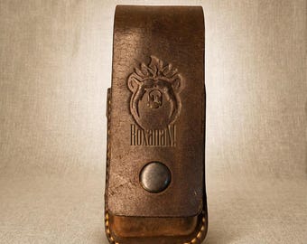 May include: A dark brown leather belt pouch featuring a vertical rectangular shape with a flap closure secured by a single metal snap button. The front is embossed with a bear head logo above the text RoxanaN! and has visible light stitching.