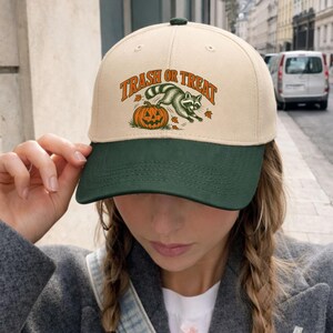 May include: A beige baseball cap with a dark green brim featuring a graphic of a raccoon jumping over a jack-o'-lantern pumpkin with the text TRASH OR TREAT in orange letters. The cap has a dark green button on the top and stitched eyelets.