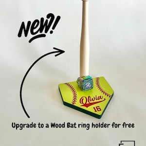 May include: A light-colored wooden baseball bat ring holder standing upright on a yellow home plate base. The base features red stitching graphics, the name Olivia, and the number 16. Text includes New!, Upgrade to a Wood Bat ring holder for free, and ELM.