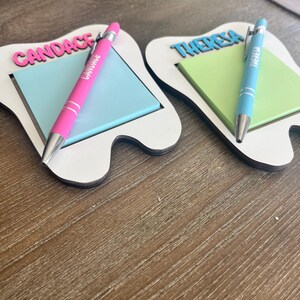 Pode incluir: Dois organizadores de mesa em forma de dente com blocos de notas e canetas. Um tem "CANDACE" em rosa, uma caneta rosa e blocos de notas azuis. O outro tem "THERESA" em azul, uma caneta azul e blocos de notas verdes. Os organizadores s&atilde;o brancos com base de madeira.