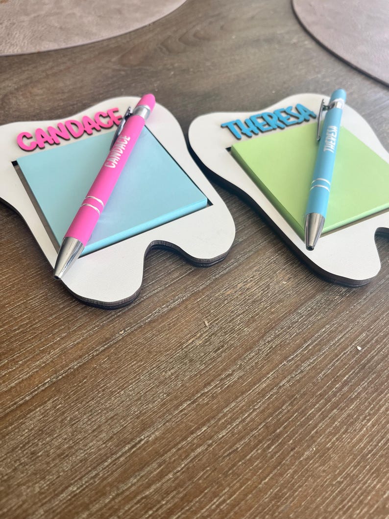 Pode incluir: Dois organizadores de mesa em forma de dente com blocos de notas e canetas. Um tem "CANDACE" em rosa, uma caneta rosa e blocos de notas azuis. O outro tem "THERESA" em azul, uma caneta azul e blocos de notas verdes. Os organizadores s&atilde;o brancos com base de madeira.