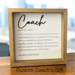 May include: A framed sign with the word "Coach" in black script. The sign includes a definition of "Coach" and the text "Custom Coach's Gift". The frame is a light brown color.