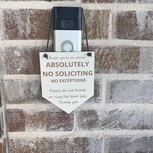 May include: A white, shield-shaped sign hanging from a silver video doorbell. The sign features black and brown text that reads: Smile, you're on camera, ABSOLUTELY NO SOLICITING NO EXCEPTIONS, Please do not knock or ring the door bell, Thank you.