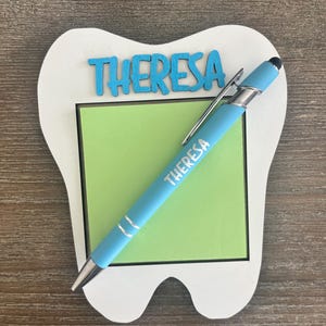 Pode incluir: Um porta-bloco em forma de dente branco com um bloco de notas verde. A palavra "THERESA" est&aacute; escrita em azul na parte superior e numa caneta azul clara. A caneta tem um clipe e uma ponta prateados.