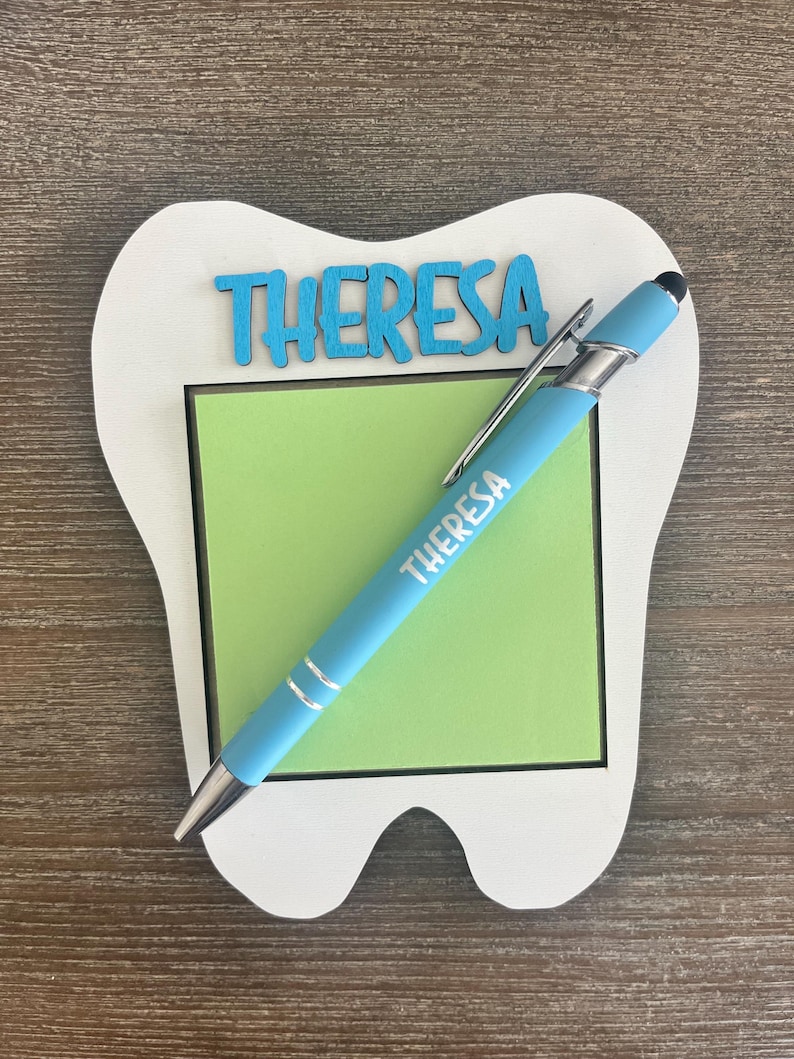 Pode incluir: Um porta-bloco em forma de dente branco com um bloco de notas verde. A palavra "THERESA" est&aacute; escrita em azul na parte superior e numa caneta azul clara. A caneta tem um clipe e uma ponta prateados.