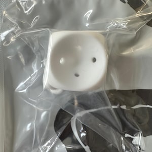 May include: A white, square-shaped product with three small holes, packaged in a clear plastic bag. The product appears to be made of a solid, smooth material. The background shows a black and white pattern.