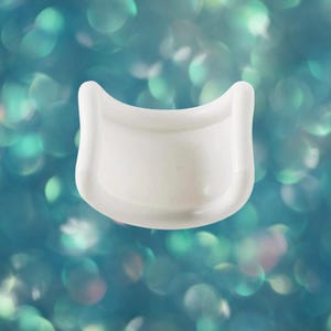 May include: A white, curved, cosmetic tool, possibly an eyelash curler, against a blurred teal background. The tool has a smooth, glossy finish and a rounded shape with a flat base. The background features out-of-focus, circular highlights.