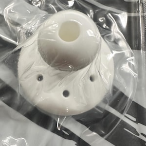 May include: A white, circular, dome-shaped object with a central opening and four smaller, evenly spaced holes around the perimeter. The object is encased in a clear, crinkled plastic film, revealing its smooth, matte, solid white surface.