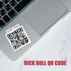 May include: A white sticker with a black QR code and the text "scan me!" is affixed to a laptop. Below the sticker, the text "RICK ROLL QR CODE" is printed in red.